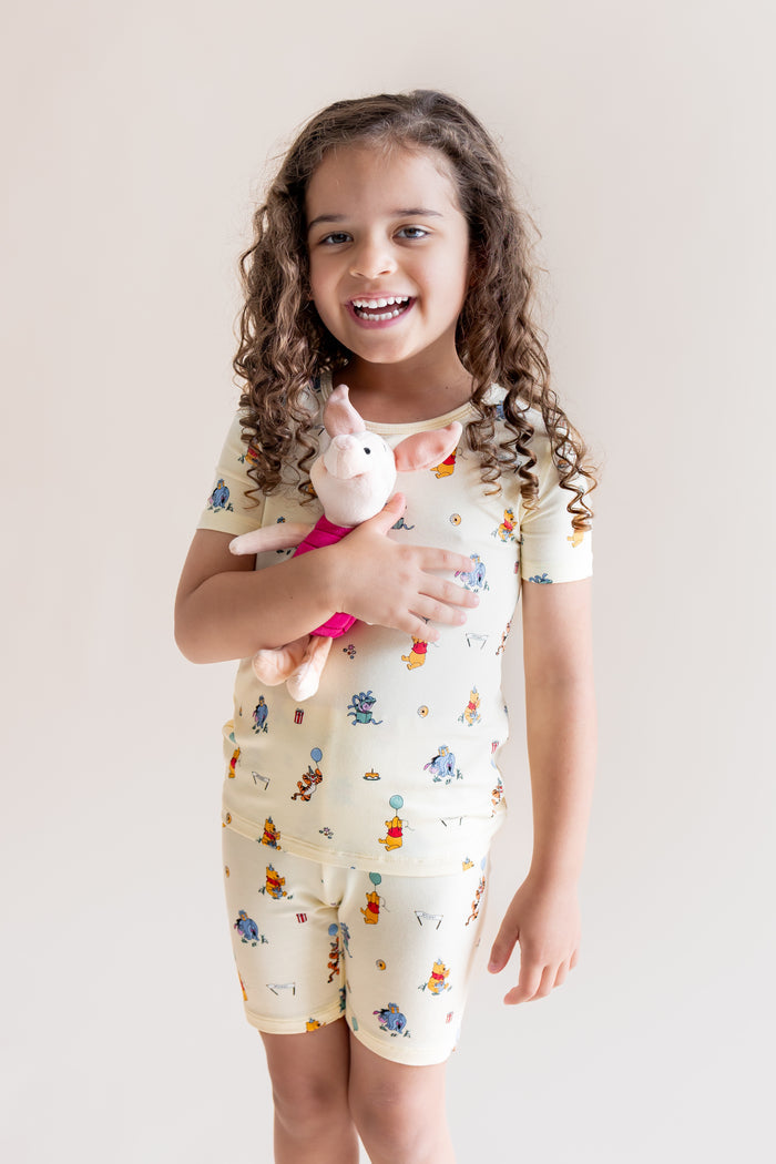 Girl holding a stuff toy wearing Short Sleeve Pajamas in Winnie the Pooh Celebration print on a cream background