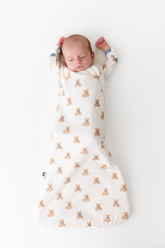 A sleeping newborn baby lying on their back, swaddled in a white long-sleeve sleep sack with a repeating pattern of small tan teddy bears. The baby's hands are raised above their head, and the scene is photographed from directly above on a clean white background.