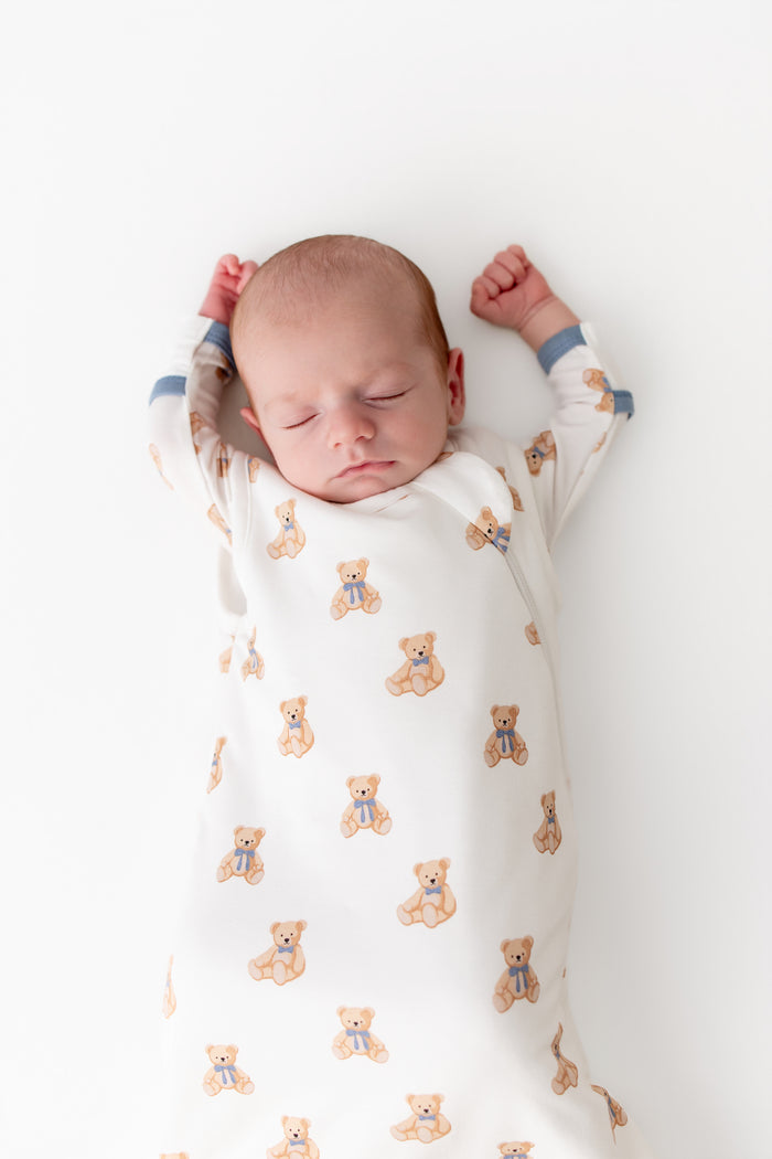 A sleeping newborn baby swaddled in a white wearable blanket with a repeating pattern of small tan teddy bears wearing blue bowties, lying on a plain white background.