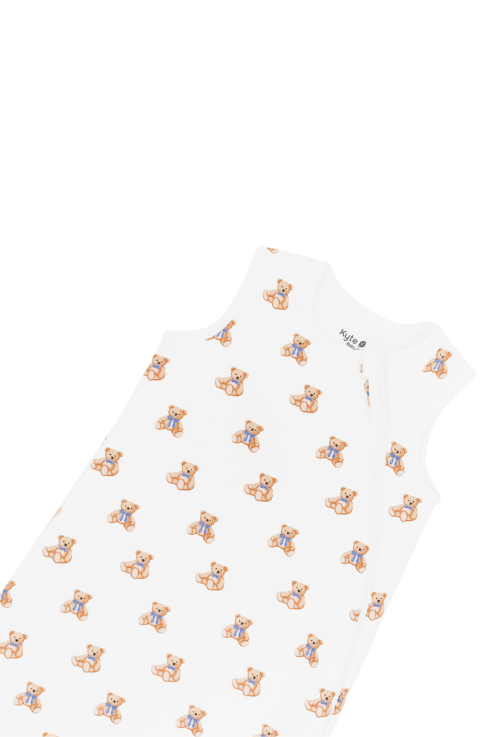 A white sleeveless baby sleep sack featuring a repeating pattern of small teddy bears wearing blue bowties, shown against a plain white background.