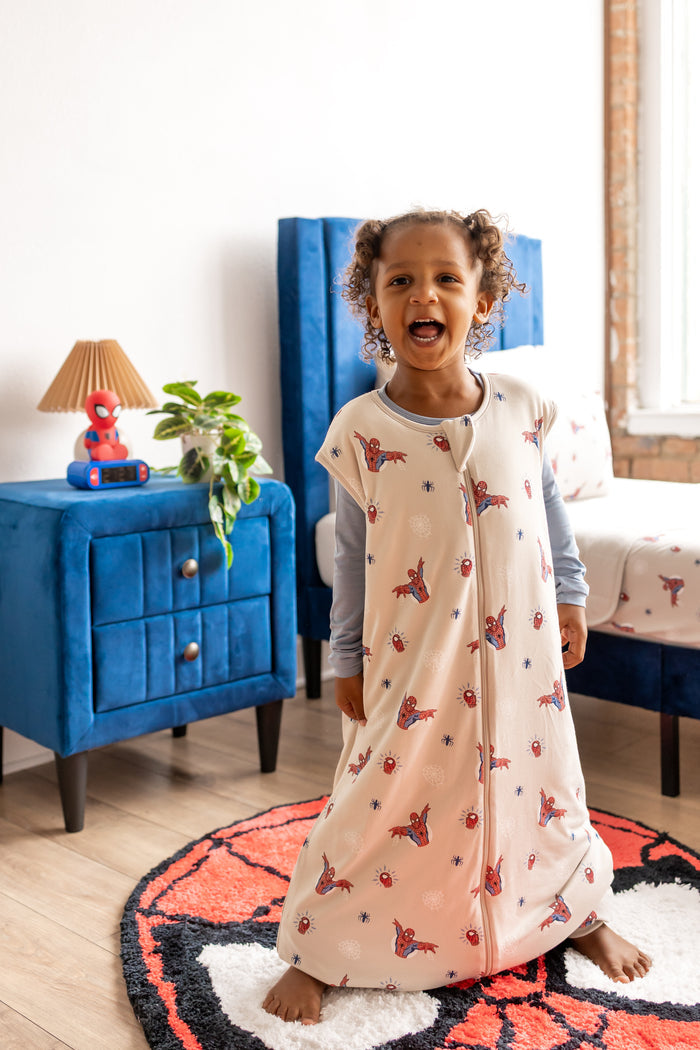 Girl standing on a spider print rug in a room with blue bed and side table wearing oat color Sleep Bag Walker with Spider-Man print