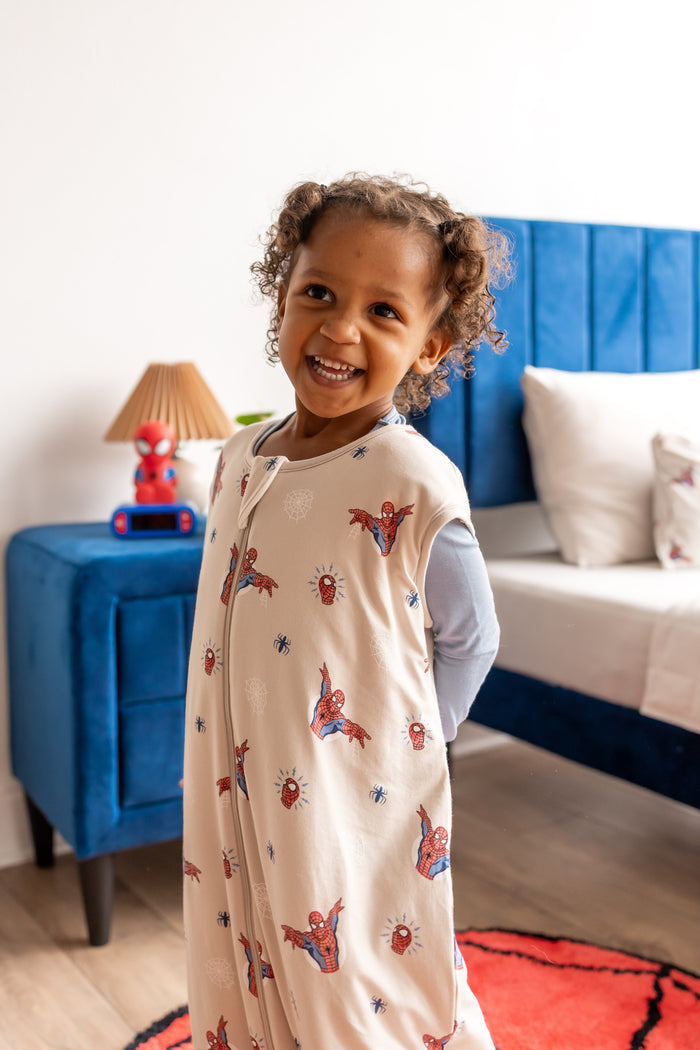 Girl standing in a room with blue bed and side table wearing an oat color Sleep Bag Walker with Spider-Man print