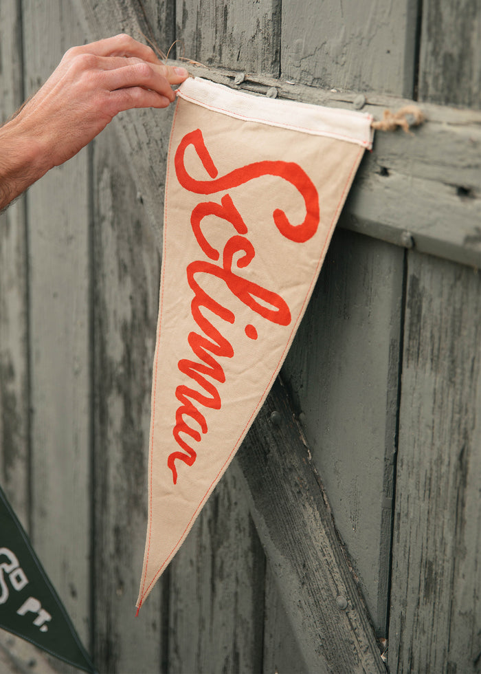 A hand hangs a beige pennant with "Selina" in red script on a rustic wooden wall, adding a vintage touch to the scene.
