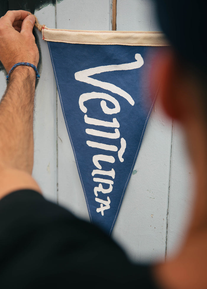 A person hangs a blue pennant with "Ventura" on a wooden wall, focusing on the text and hand.