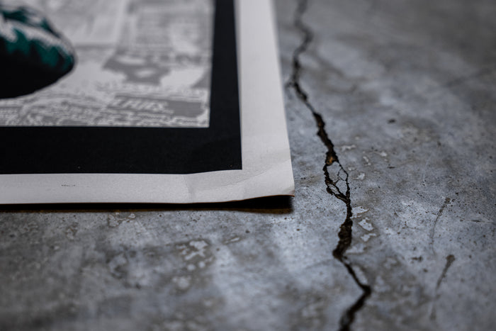 A close-up of the corner of a damaged 18x24 Bane poster lying on a cracked concrete floor, showing newspaper-like text and a dark border.