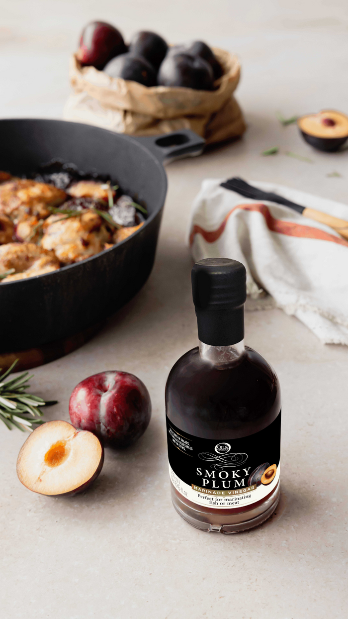 Bottle of Smoky Plum syrup on a table with plums and a skillet in the background