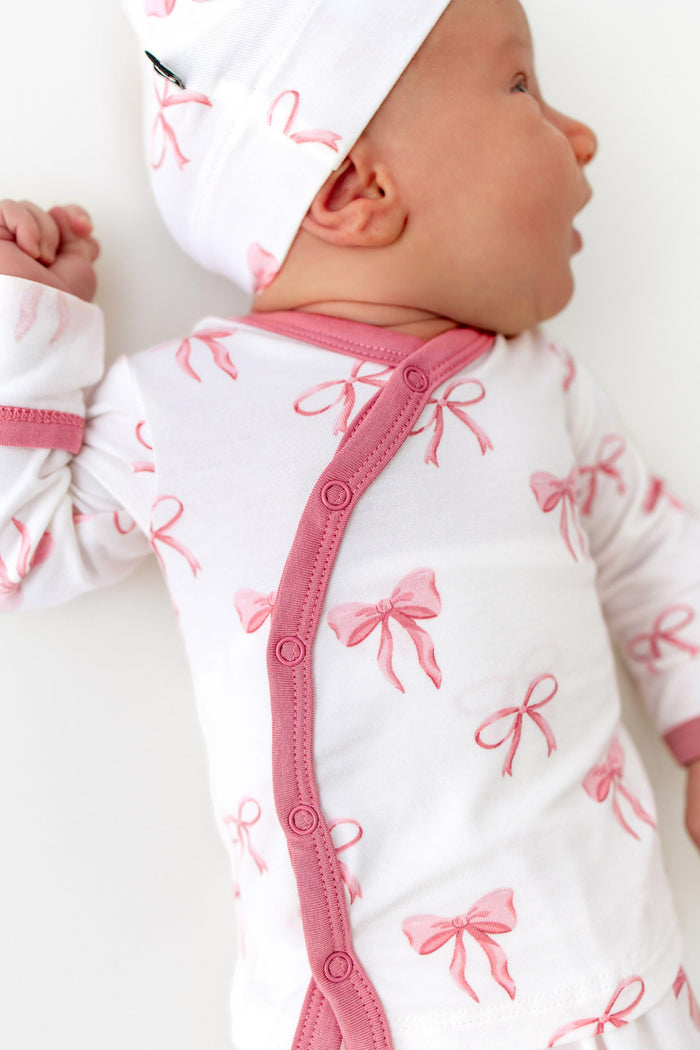 Baby wearing a white outfit with pink bows and a matching hat on a white background