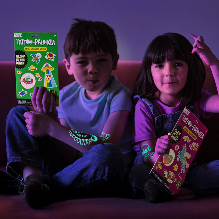 Two kids in darkly lit room holding sets of OOLY Tattoo-Palooza Glow in the Dark Temporary Tattoos