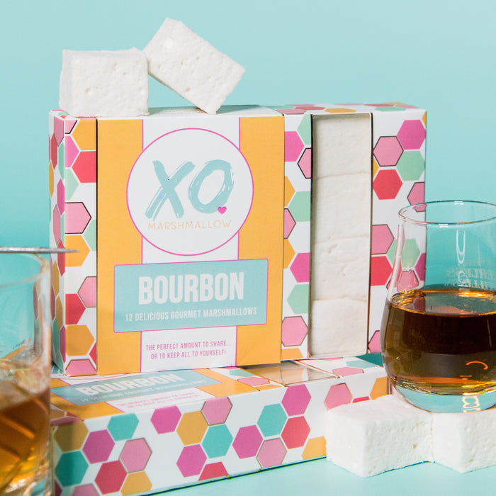 A box of gourmet xo marshmallow bourbon marshmallows against a blue background with a glass of bourbon neat next to it