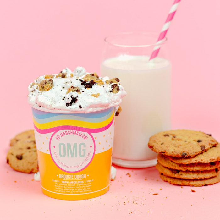 A pint of XO Marshmallow Gourmet Brookie Dough Marshmallow fluff with a glass of milk with a pink striped straw in the background. There is a stack of chocolate chip cookies next to it.