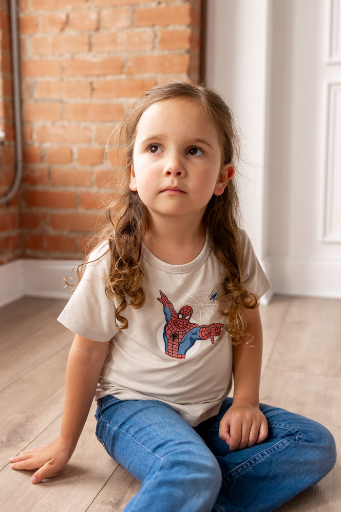 Girl sitting on floor in front of a brick wall wearing oat color Toddler Basic Tee with Spider-Man print in center over blue jeans
