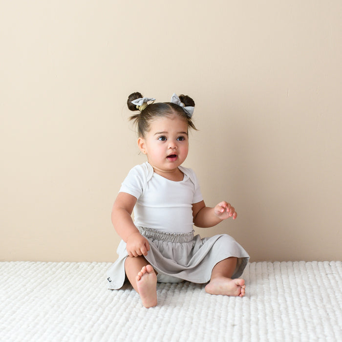 Toddler sitting on the floor while wearing a Toddler Skort in Oat, Short Sleeve Bodysuit in Cloud and pigtails with Bows in Oat