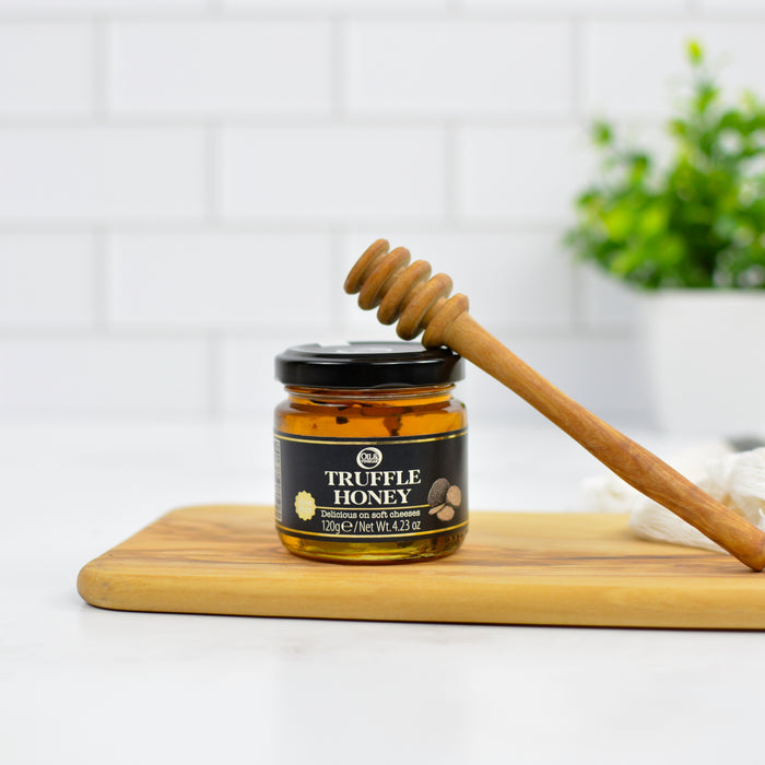 Jar of truffle honey with a wooden dipper on a wooden board against a white tiled wall.