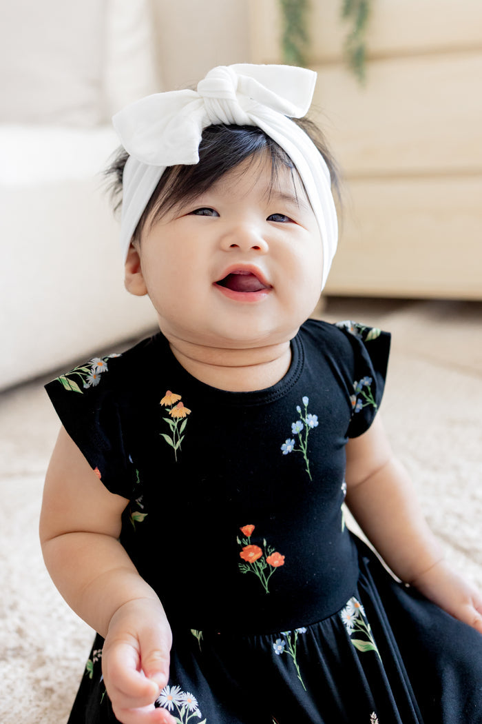 Baby smiling in Twirl Bodysuit Dress in Midnight Wildflower print wearing a white head bow on a white carpet