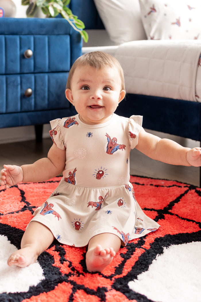 Baby wearing oat color Twirl Bodysuit Dress with Spider-Man print sitting on a spider man print rug