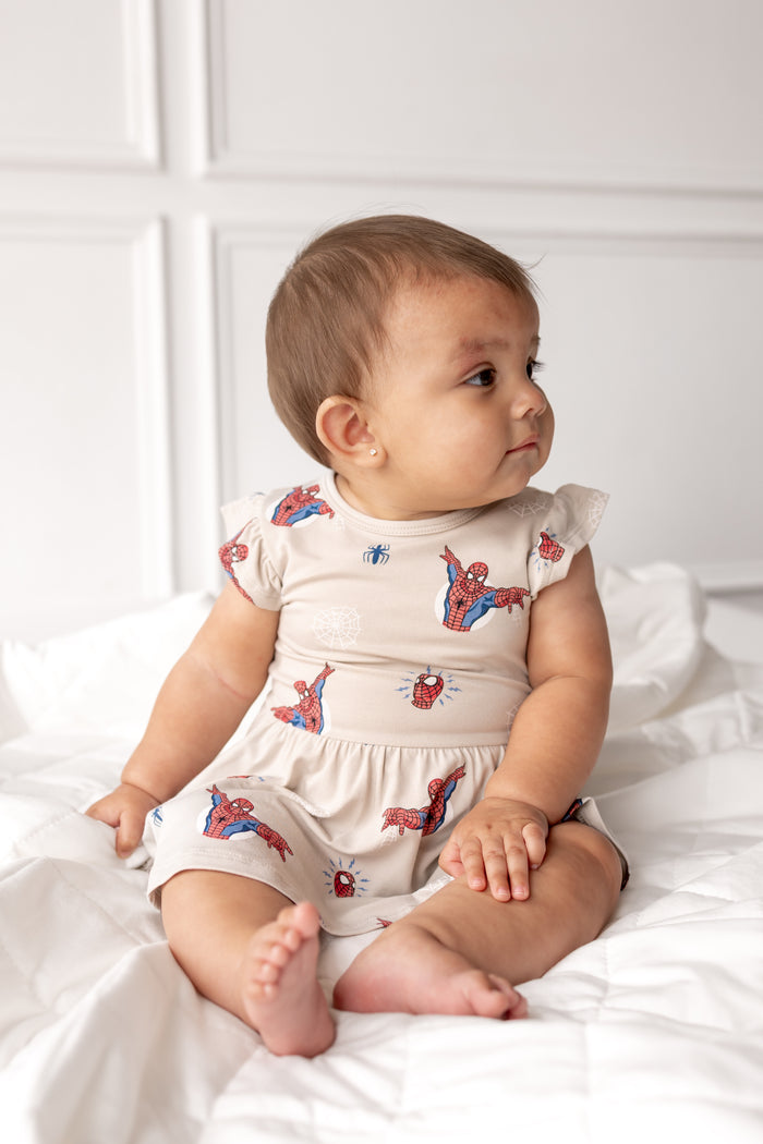 Baby wearing oat color Twirl Bodysuit Dress with Spider-Man print sitting on a white bed