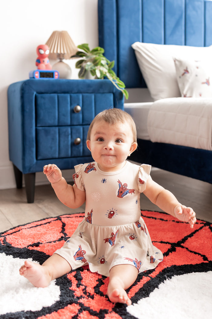 Baby wearing oat color Twirl Bodysuit Dress with Spider-Man print sitting on a spider man print rug in a room with blue bed and side table