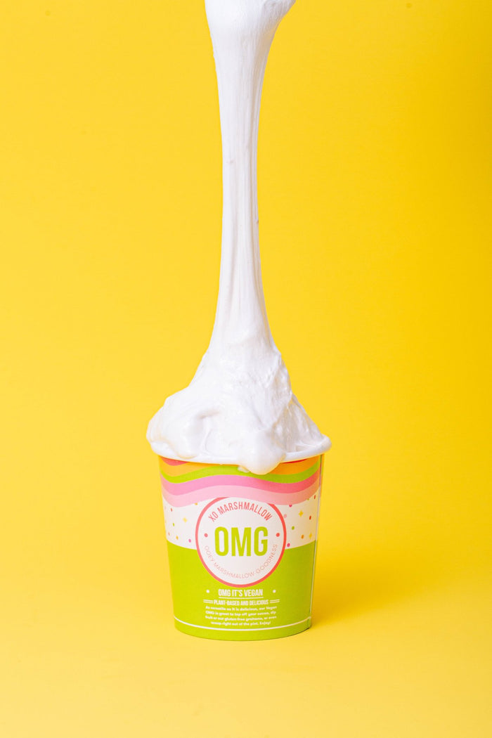 A pint of vanilla flavored vegan marshmallow fluff with a long stretchy pull of marshmallow creme coming out of the top connecting to the spoon at the top of the photo. All of this is against a bright yellow background.