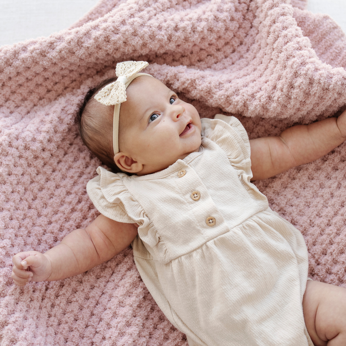 Baby girl in a cream outfit lays on a pink waffle knit receiving baby blanket.