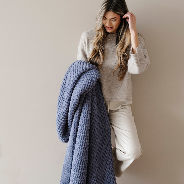 A woman is holding a blue waffle knit blanket that is a Saranoni blanket. This soft blanket can be used for relaxing, giving as a comfort gift, or home decor. Enjoy this durable cozy throw blanket for years to come!