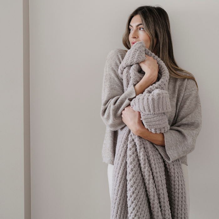 A woman looks in the distance and she is holding a light tan waffle knit blanket. This Saranoni blanket is a throw blanket and is a beautiful home decor piece. It can also be used for relaxing or gifting.