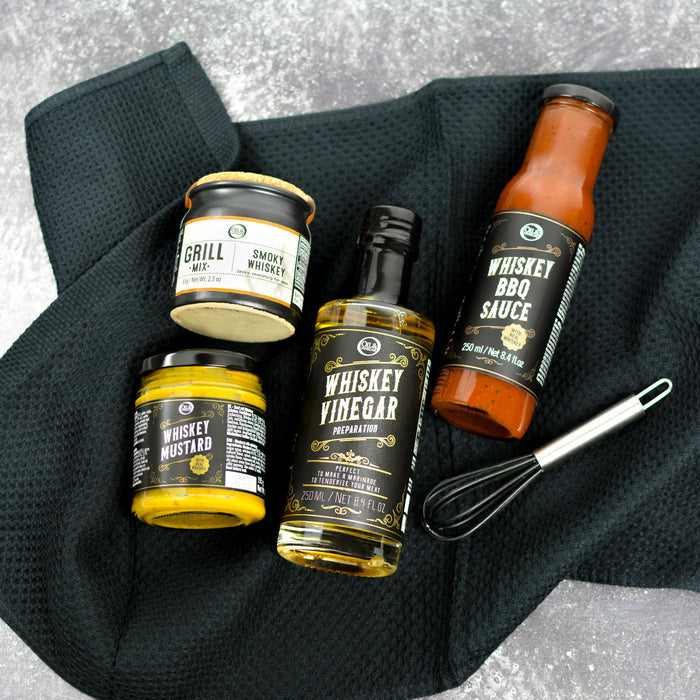 A collection of whiskey-themed food items including whiskey BBQ sauce, whiskey mustard, whiskey vinegar, and a smoky whiskey grill mix, presented with a black mini whisk on a textured black surface.