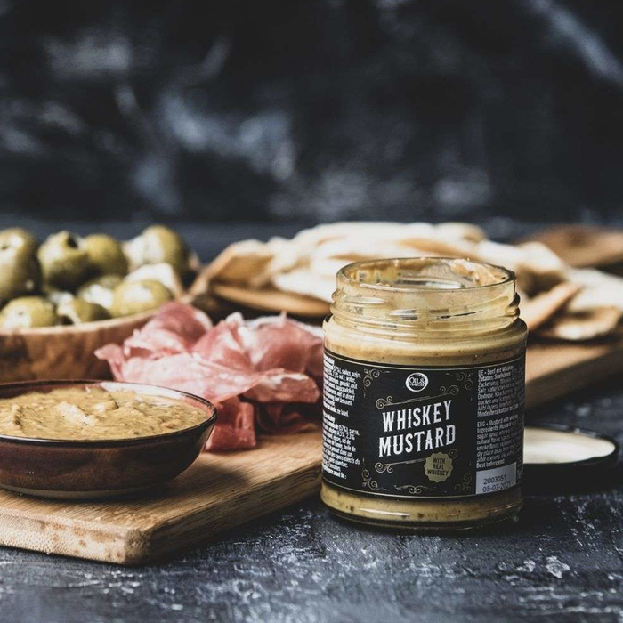 jar of Whiskey Mustard wit ha black label on a black background next to a charcuterie board with crackers, olives, cured meats, and a dish of the Whiskey Mustard