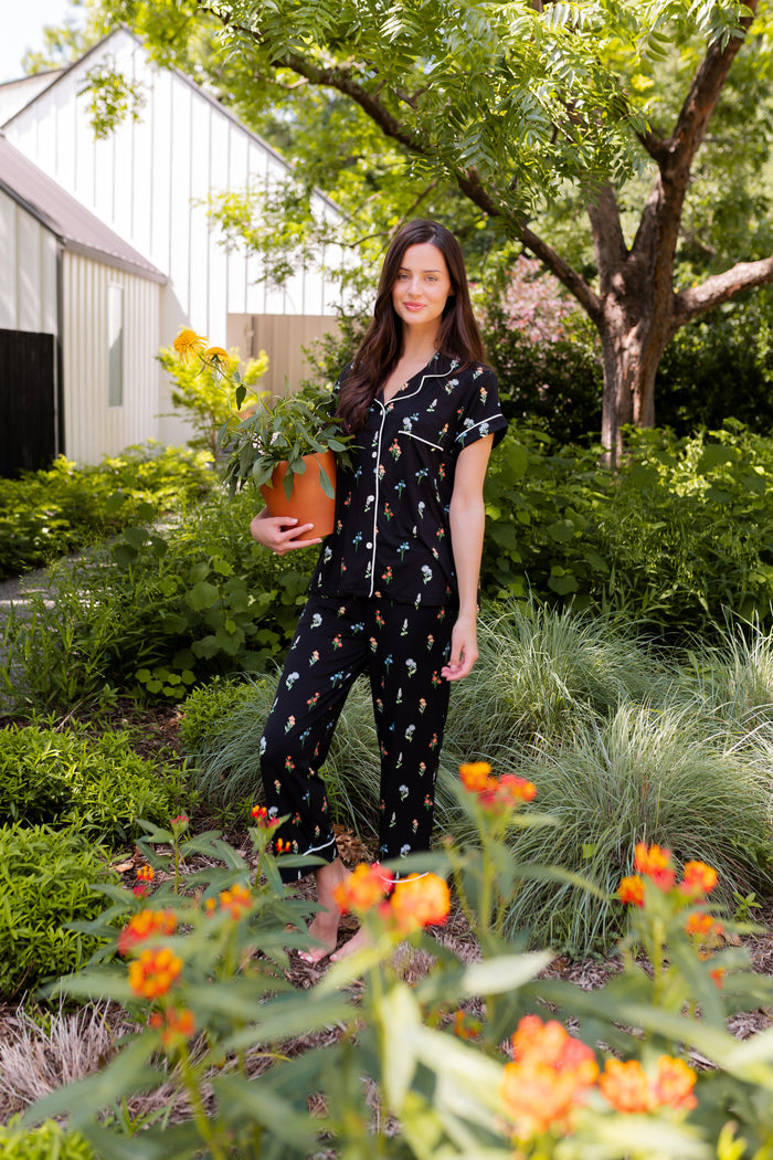Model standing outdoors surrounded by trees holding a pot wearing Women's Short Sleeve Cropped Pajama Set in Midnight Wildflower print