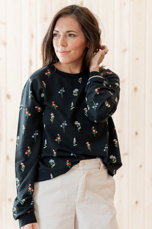 A woman wearing a black crewneck sweatshirt with a delicate multicolor floral print, paired with beige pants, standing against a light wood-paneled wall.