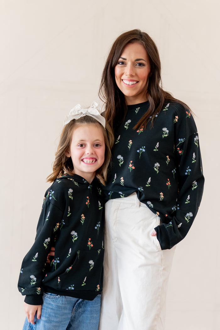 A woman and a young girl wearing matching black crewneck sweatshirts with a small multicolor floral print, posed against a plain beige studio background.