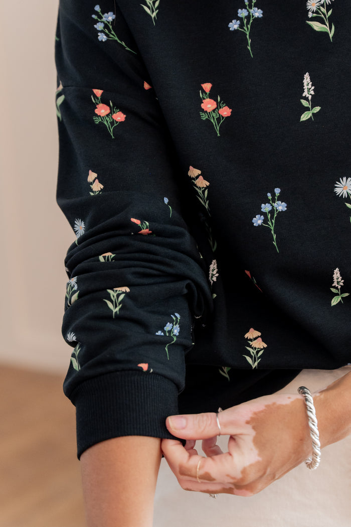 Close-up of a person wearing a black long-sleeve sweatshirt with a delicate multicolor floral print, showing the ribbed cuff being adjusted.