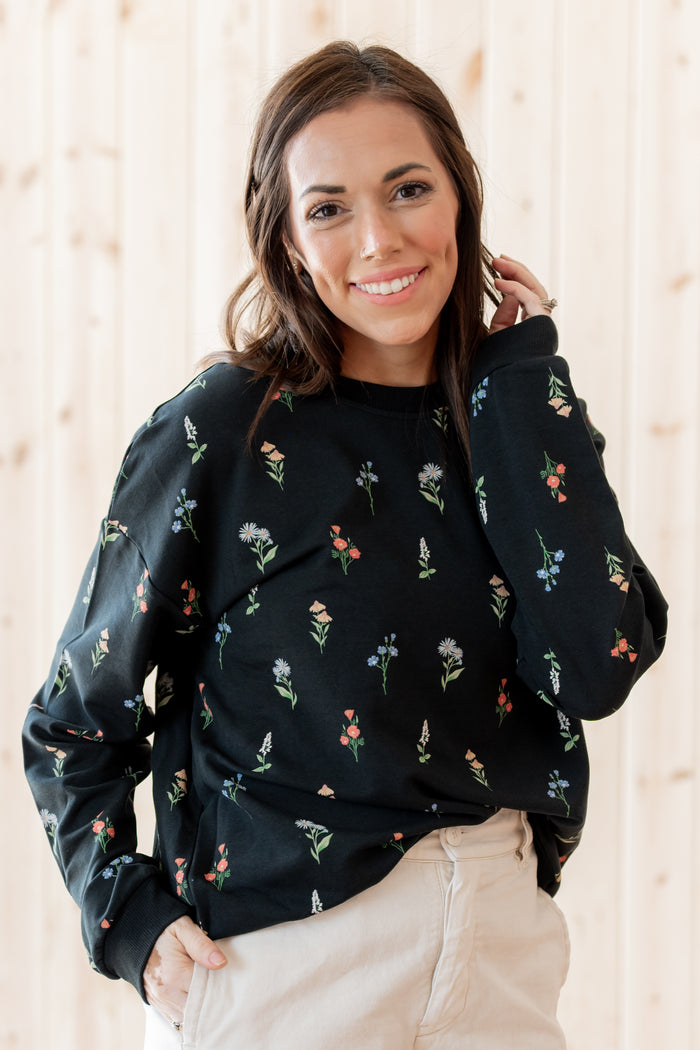 A woman wearing a black crewneck sweatshirt with a small multicolor floral print, paired with light beige pants, standing against a light wood-paneled wall.
