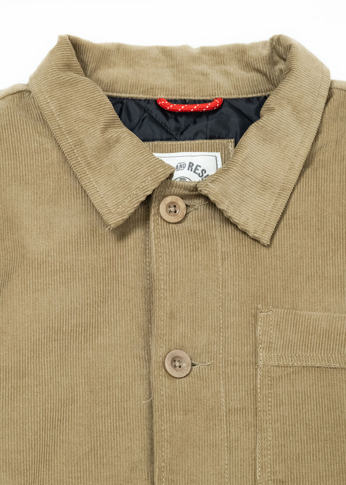Tan corduroy jacket with buttons, collar, and chest pocket. Red loop inside collar. Close-up view, highlighting texture.