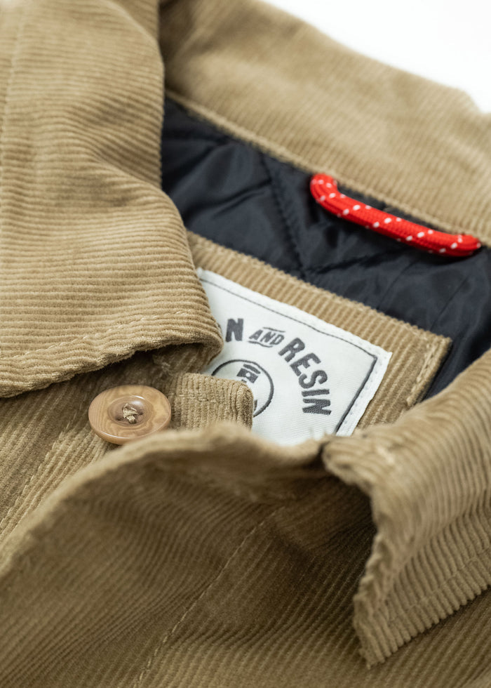 Close-up of a tan corduroy jacket with a button, visible label, and red loop. The label reads "Iron and Resin."