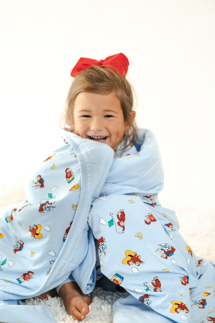 Girl wrapped in Youth Blanket in Curious George 2.5 print wearing red bow on head sitting on a white carpet