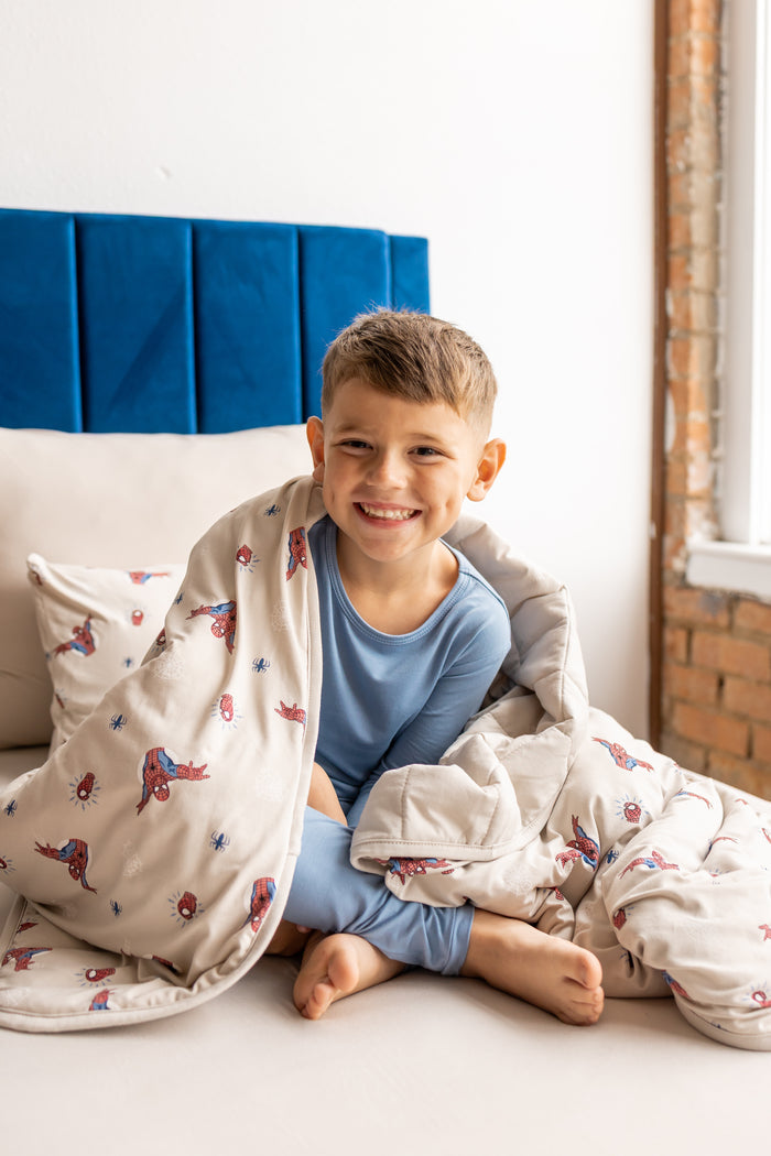 Boy sitting on a bed with oat color sheets and pillow wrapped in oat color Youth Blanket with Spider-Man print wearing blue pajamas