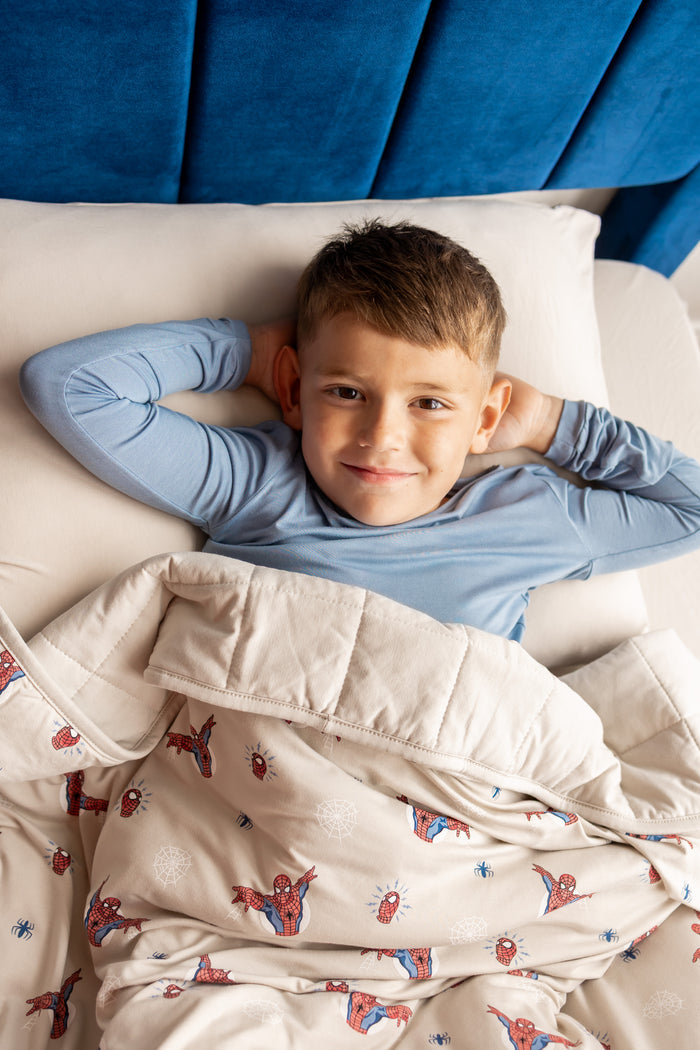 Boy lying on bed covered under an oat color Youth Blanket with Spider-Man print wearing blue shirt