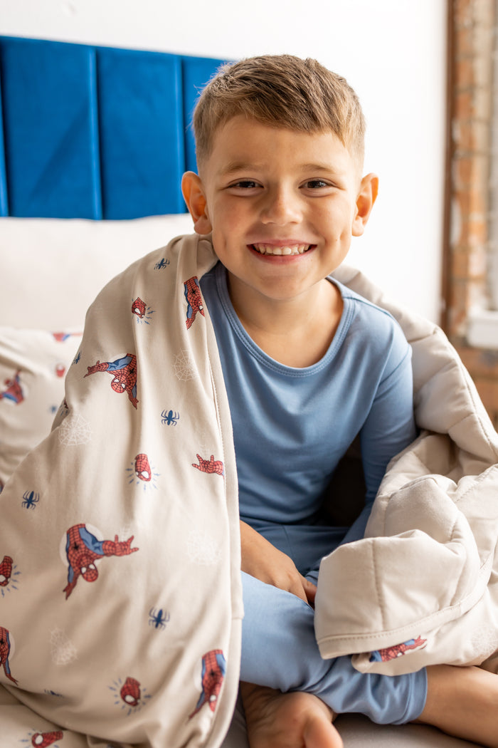 Boy smiling on bed wrapped in oat color Youth Blanket with Spider-Man print wearing blue pajamas