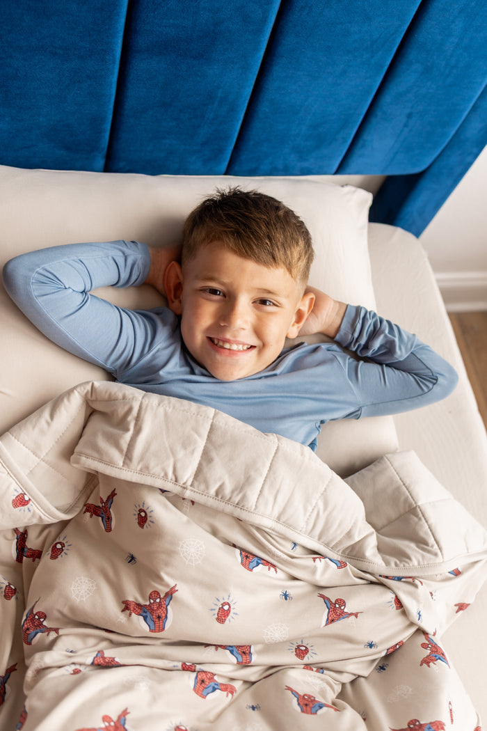Boy lying on a bed under an oat color Youth Blanket with Spider-Man print wearing blue pajamas