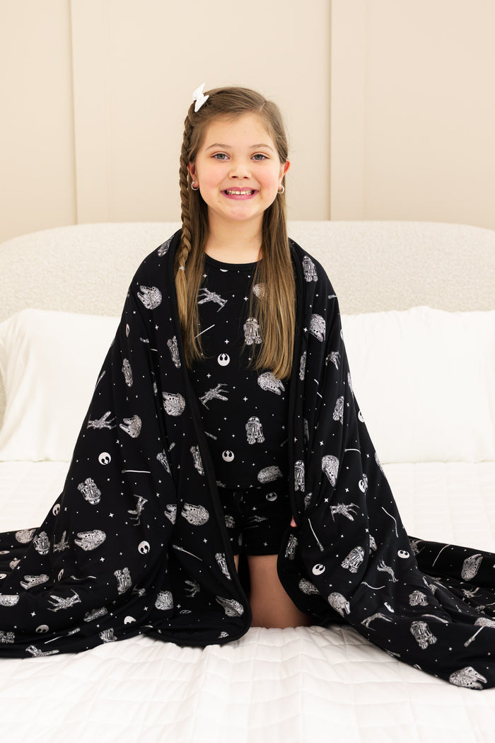 Girl sitting on white bed wrapped with Youth Blanket in STAR WARS™ 2.5 print wearing similar print short sleeve pajama set