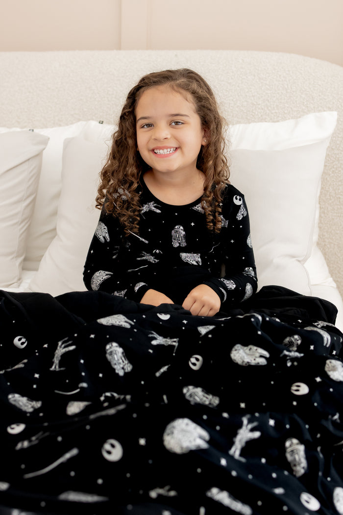 Girl sitting on bed covered under Youth Blanket in STAR WARS™ 2.5 print wearing similar print long sleeve pajama set