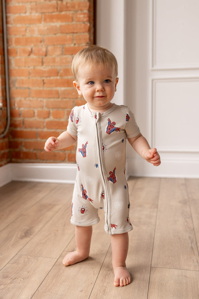 Toddler wearing oat color Zipper Shortall Romper with Spider-Man print standing in front of a brick wall