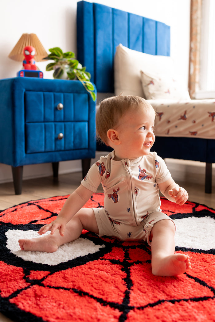 Toddler wearing oat color Zipper Shortall Romper with Spider-Man print sitting on a spider man print rug in a room