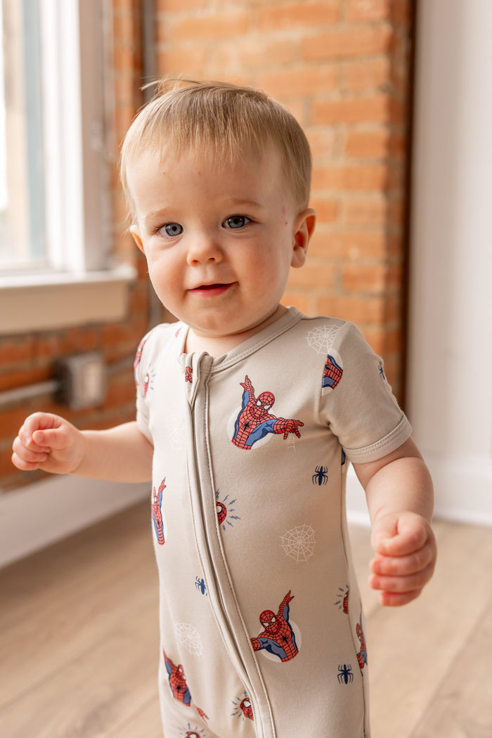 Toddler wearing oat color Zipper Shortall Romper with Spider-Man print standing in front of a brick wall